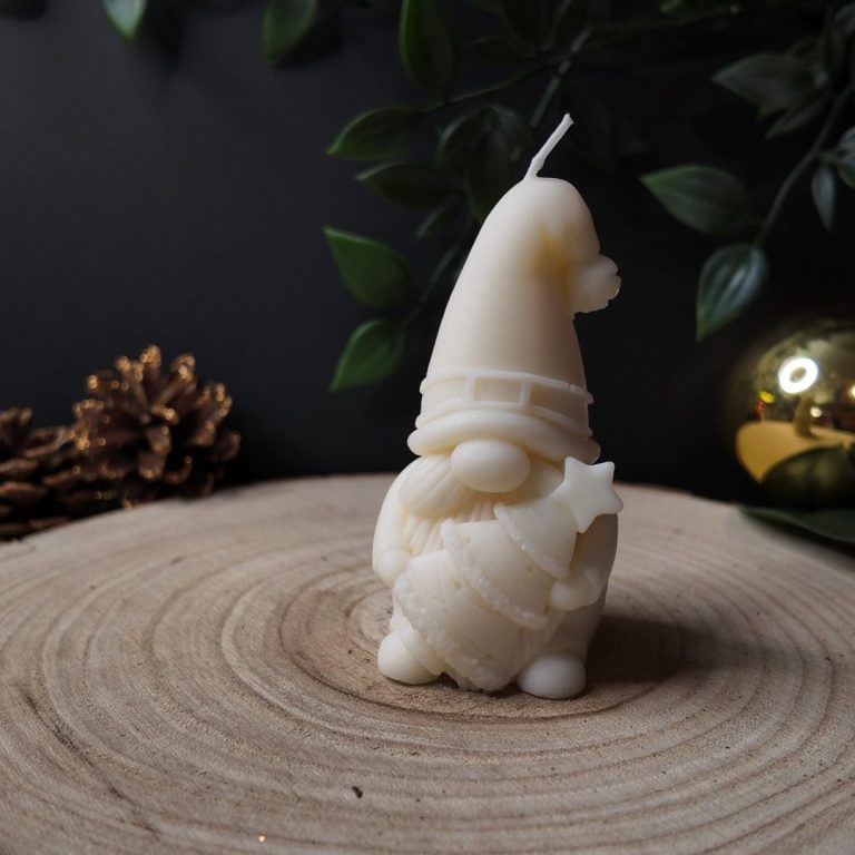 Weihnachtswichtel Kerze aus Naturwachs Eine weiße Kerze in Form eines Gnoms mit einem Hut, umgeben von Tannenzweigen und einer Weihnachtskugel.